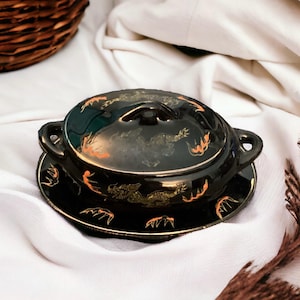 May include: Black ceramic serving bowl with lid and saucer. The bowl and lid feature a dragon design in gold and orange. The bowl has two handles and sits on a matching saucer with a gold trim. The set is on a white cloth.