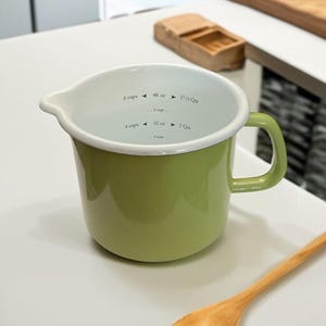 May include: A green enamel measuring cup with a white rim and handle. The interior has black markings for cups, ounces, and quarts. A wooden spoon rests nearby. Kitchenware.