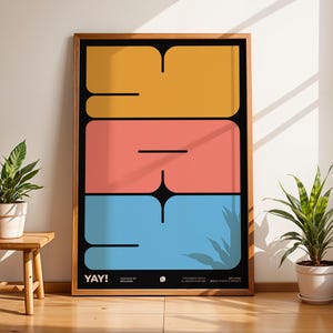 K&ouml;nnte beinhalten: Ein gerahmter Kunstdruck mit dem Wort "YAY!" in einem kr&auml;ftigen, farbenfrohen Design. Das Kunstwerk zeigt gro&szlig;e, gestapelte Formen in Gelb, Koralle und Blau vor einem schwarzen Hintergrund. Der Druck wird in einem Raum mit Pflanzen und nat&uuml;rlichem Licht ausgestellt.