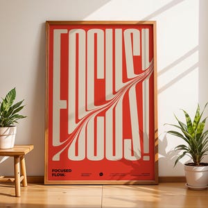 Puede incluir: Un póster enmarcado con la palabra "FOCUS!" en letras grandes y estilizadas de color blanco roto sobre un fondo rojo. El póster está en un marco de madera e incluye el texto "FOCUSED FLOW." Se ven dos plantas en macetas.