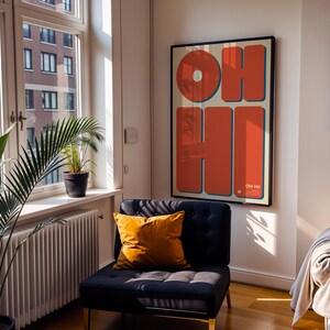 May include: A framed art print with the words "OH HI" in large, orange letters with blue outlines. The artwork hangs on a white wall above a dark blue armchair with a yellow pillow. A window with plants and a radiator is to the left.