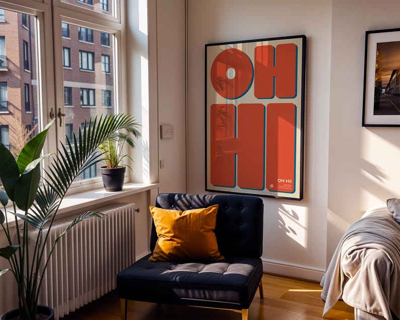 May include: A framed art print with the words "OH HI" in large, orange letters with blue outlines. The artwork hangs on a white wall above a dark blue armchair with a yellow pillow. A window with plants and a radiator is to the left.