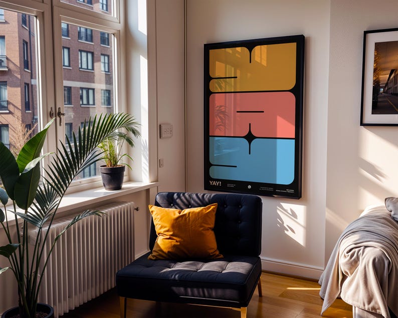 K&ouml;nnte beinhalten: Ein gerahmter Kunstdruck mit schwarzem Rahmen, der drei gestapelte, farbige rechteckige Formen in Gelb, Koralle und Blau zeigt. Das Wort "YAY!" steht unten. Ein marineblauer Stuhl mit einem orangefarbenen Kissen steht daneben.