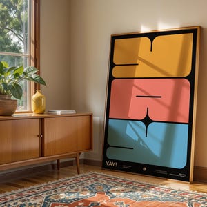 K&ouml;nnte beinhalten: Ein gerahmter Kunstdruck mit einem kr&auml;ftigen, geometrischen Design in Gelb, Koralle und Blau auf schwarzem Hintergrund. Das Wort "YAY!" steht unten. Der Druck lehnt an einer Wand neben einem Holzschrank und einem Fenster.