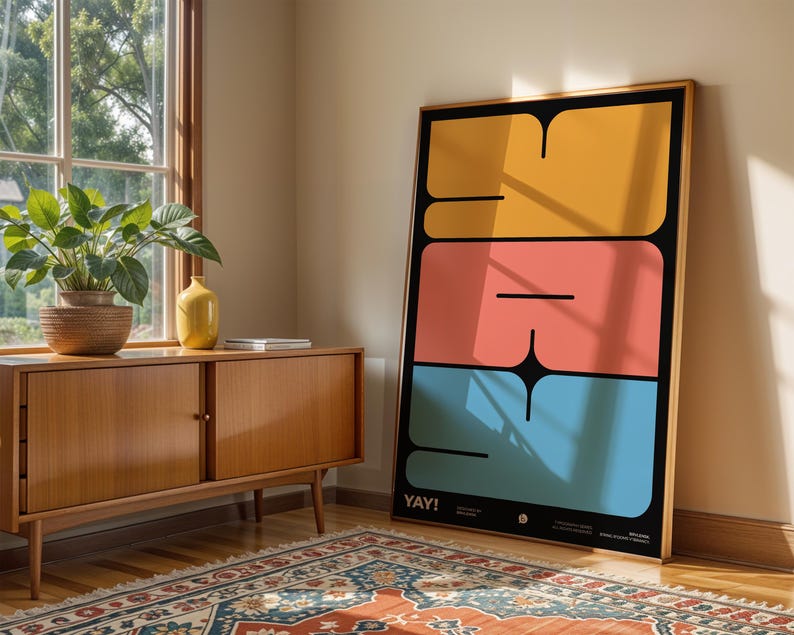 K&ouml;nnte beinhalten: Ein gerahmter Kunstdruck mit einem kr&auml;ftigen, geometrischen Design in Gelb, Koralle und Blau auf schwarzem Hintergrund. Das Wort "YAY!" steht unten. Der Druck lehnt an einer Wand neben einem Holzschrank und einem Fenster.