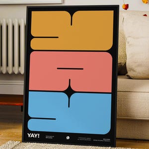K&ouml;nnte beinhalten: Ein gerahmter Kunstdruck mit dem Wort "YAY!" in gro&szlig;en, farbenfrohen, abstrakten Formen. Das Design zeigt einen schwarzen Hintergrund mit gestapelten Bl&ouml;cken in Orange, Koralle und Blau. Der Druck wird in einem Wohnzimmer gezeigt.