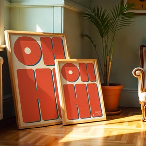 May include: Two framed art prints with the words "OH HI" in large, orange, retro-style lettering. The letters have a blue outline and are set against a cream background. The frames are light wood. One print is larger than the other.