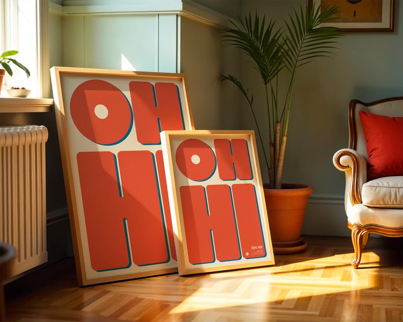 May include: Two framed art prints with the words "OH HI" in large, orange, retro-style lettering. The letters have a blue outline and are set against a cream background. The frames are light wood. One print is larger than the other.