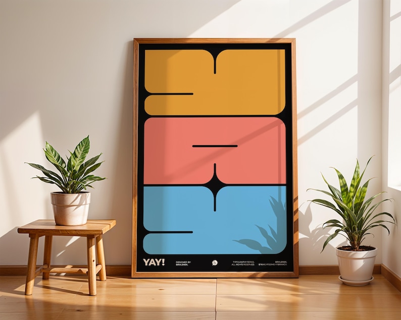 K&ouml;nnte beinhalten: Ein gerahmter Kunstdruck mit dem Wort "YAY!" in einem kr&auml;ftigen, farbenfrohen Design. Das Kunstwerk zeigt gro&szlig;e, gestapelte Formen in Gelb, Koralle und Blau vor einem schwarzen Hintergrund. Der Druck wird in einem Raum mit Pflanzen und nat&uuml;rlichem Licht ausgestellt.