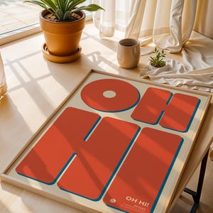 May include: A framed art print with the words "OH HI!" in large, bold, orange letters with blue outlines. The artwork is on a cream-colored background. The print is displayed on a wooden table near a window.