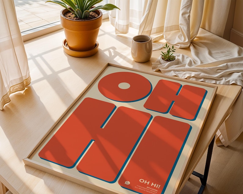 May include: A framed art print with the words "OH HI!" in large, bold, orange letters with blue outlines. The artwork is on a cream-colored background. The print is displayed on a wooden table near a window.