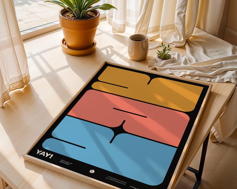 K&ouml;nnte beinhalten: Ein gerahmter Kunstdruck mit modernem Design. Er zeigt drei horizontale Bl&ouml;cke in Gelb, Koralle und Blau auf schwarzem Hintergrund. Das Wort "YAY!" ist wei&szlig; gedruckt. Der Druck wird auf einem Holztisch in der N&auml;he eines Fensters pr&auml;sentiert.