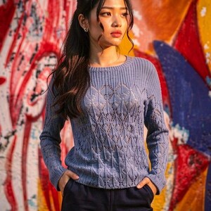 May include: A light blue, long-sleeved sweater with a detailed openwork pattern. The sweater is paired with dark blue pants. The background features colorful graffiti art.