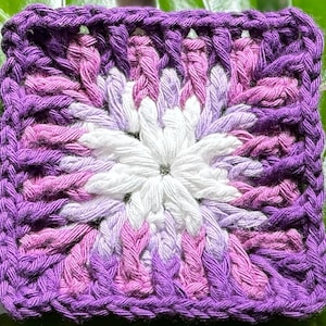 Könnte beinhalten: Ein quadratisches, handgefertigtes Häkelstück mit einem floralen Design. Die Mitte ist weiß, umgeben von Rosa- und Lilatönen, mit einem tiefvioletten Rand. Das Garn ist dicht gewebt und erzeugt ein strukturiertes Aussehen.