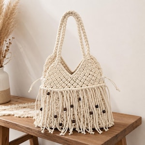 May include: An off-white macrame tote bag featuring a woven diamond pattern, two braided shoulder straps, and a drawstring closure. The lower section is adorned with long fringe strands accented by small, dark brown wooden beads throughout.