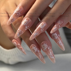 May include: Long, almond-shaped artificial nails with a nude base and delicate white floral designs. The nails feature a gradient effect, transitioning from a darker nude at the base to a lighter shade at the tips. Text on the image reads "Chae By Her Etsy Where Art Meets Elegance."