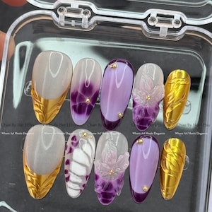May include: A set of ten artificial nails in a clear plastic case. The nails feature various designs, including gold leaf accents, purple ombre, floral embellishments, and gold studs. The text "Chae By Her | Etsy" and "Where Art Meets Elegance" are visible.