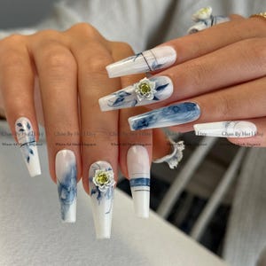 Op de afbeelding: Een set lange, vierkante kunstnagels. De nagels hebben een witte basis met blauwe bloemmotieven, aquareleffecten en 3D-bloemaccenten. De tekst "Chae By Her | Etsy" is zichtbaar op sommige nagels.