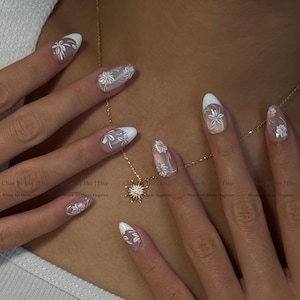 May include: Close-up of manicured nails with white floral designs and white tips. A gold necklace with a sunburst pendant is also visible. The nails are decorated with delicate white floral patterns. The text "Chae By Her | Etsy" is visible.