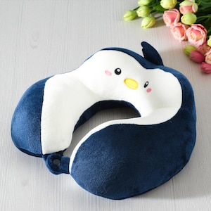 Op de afbeelding: Een marineblauw en wit pluche reiskussen in de vorm van een pinguïn. Het U-vormige kussen heeft een knoopsluiting. De pinguïn heeft een gele snavel, roze wangen en zwarte ogen. Het kussen is ontworpen voor nekondersteuning.