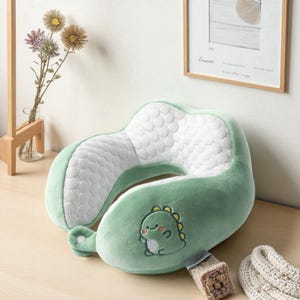 May include: A green travel neck pillow with a dinosaur design. The pillow has a U-shape with a white patterned center and a green exterior. A cartoon dinosaur is embroidered on the front. The pillow is designed for comfort and support during travel.