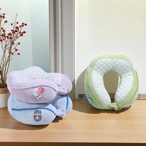 May include: Three travel neck pillows in pastel colors. One is light green, another is lavender, and the third is light blue. Each pillow has a different embroidered cartoon animal. The pillows are on a wooden surface.