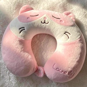 May include: A soft, plush U-shaped neck pillow in light pink and white, designed to resemble a cat face with closed eyes, a small nose, and whiskers. The word Lovely is embroidered in cursive script on the lower right side of the pillow.