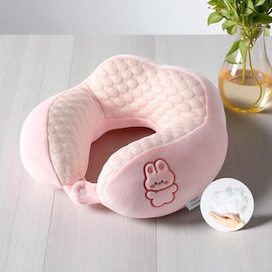 Pode incluir: Uma almofada de pescoço rosa claro para viagem com um design de coelho bordado. A almofada tem um centro acolchoado e um exterior macio e felpudo. Uma pequena etiqueta está anexada. Projetada para conforto e suporte durante a viagem.