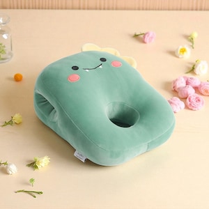 Peut inclure: Un chauffe-mains en peluche vert menthe en forme de dinosaure avec un visage souriant, des joues roses et des accents jaunes. Le chauffe-mains a un trou au centre et deux ouvertures latérales pour les mains. Une petite étiquette est visible sur le côté.