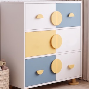 May include: A three-drawer chest featuring a white frame with alternating pastel blue and yellow drawer fronts. The drawers are accented with circular and semi-circular yellow wooden knobs. The unit is supported by four short, cylindrical yellow legs.
