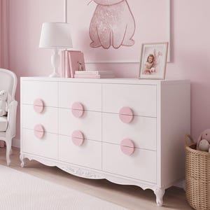 May include: A white dresser with nine drawers featuring round pink knobs, positioned in a room with pink walls. A white table lamp, books, and a framed picture sit on top. A white armchair with plush toys and a woven basket are nearby.