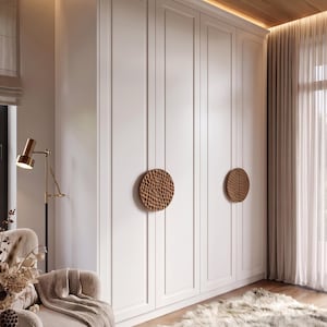 May include: A tall white wardrobe featuring four vertical panels and two large, circular, textured wooden door handles. The cabinet has a clean, flat-front design with recessed detailing on each door, providing a structured appearance for storage.