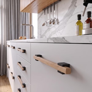 Puede incluir: Un tirador de cajón de cocina que presenta una barra horizontal de madera clara con soportes de montaje cuadrados de metal negro en cada extremo. El tirador está fijado a una puerta de armario blanca mate con veta de madera visible.