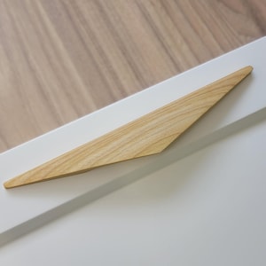 May include: A light-coloured wooden cabinet pull with a triangular design is mounted on a white cabinet door. The door is set against a light brown wooden surface.