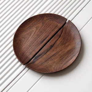 May include: A circular dark brown wooden plate featuring a distinct diagonal split that divides the surface into two separate pieces. The wood grain is visible across the smooth, polished surface of both sections of the plate.