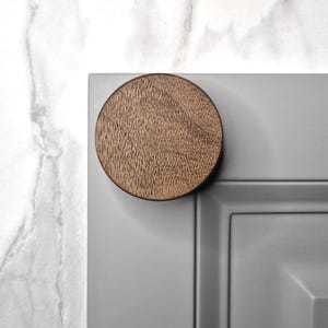 May include: A round, wooden cabinet knob with a natural wood grain finish is attached to a gray cabinet door. The knob is centered on the door's edge, providing a simple, modern design element. The background is a white marble surface.
