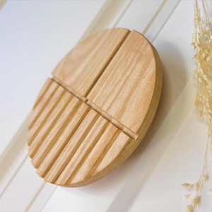 Könnte beinhalten: Eine hellfarbene, ovale Seifenschale aus Holz mit gerilltem Design auf einer Seite. Die Schale ist in vier Abschnitte unterteilt. Getrocknete, beige dekorative Stiele liegen neben der Schale auf einer weißen Oberfläche.