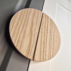 May include: A round, light-colored wooden door handle, split into two halves with a vertical groove. The handle is mounted on a white door, with a gray door frame visible in the background.