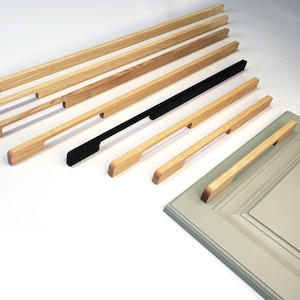 May include: A collection of linear cabinet pulls in varying lengths, featuring natural light wood grain finishes and one solid black matte finish. These long, slender handles have a recessed grip design and are displayed alongside a cabinet door.
