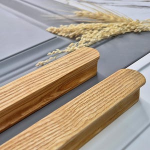 May include: Two wooden cabinet pulls with a natural wood grain finish. The pulls are rectangular with rounded ends. The background features a grey cabinet door and decorative wheat stalks.