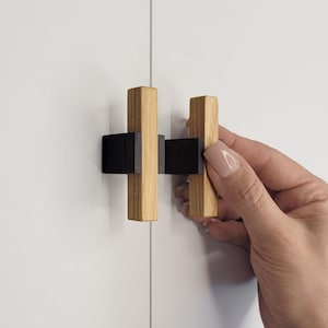 May include: A pair of wooden cabinet pulls. Each pull features a natural wood grain vertical post with a black horizontal accent. The pulls are mounted on a white cabinet door. The cabinet hardware is being held by a hand.