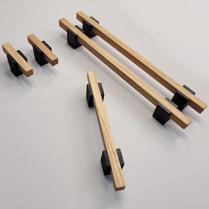 Może przedstawiać: Zestaw drewnianych uchwytów i gałek do szafek. Uchwyty mają naturalne wykończenie słojów drewna i są montowane na czarnych prostokątnych podstawach. Uchwyty są prostokątne i różnej wielkości.