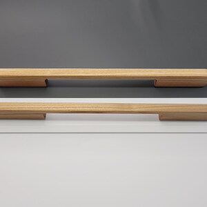 May include: Two wooden cabinet pulls with a natural finish. The pulls are rectangular with a slightly rounded top and a recessed area on each end for mounting. The background is a mix of white and gray.