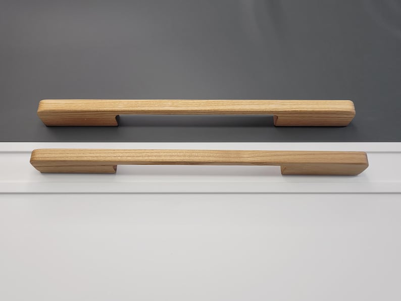 May include: Two wooden cabinet pulls with a natural finish. The pulls are rectangular with a slightly rounded top and a recessed area on each end for mounting. The background is a mix of white and gray.