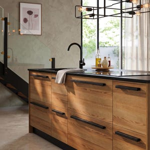 May include: A kitchen island featuring light wood cabinets with horizontal black metal handles and a black countertop. A black faucet, a light-colored cloth, and a wooden tray with glass bottles are placed on the surface under a black metal light fixture.