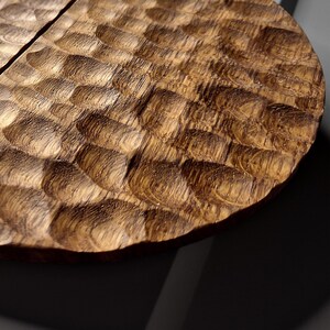 Può includere: Un sottobicchiere circolare in legno caratterizzato da una texture intagliata a mano a forma di scaglie su tutta la superficie. Il legno presenta una ricca tonalità marrone medio con venature visibili e una serie di rientranze concave.