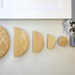 May include: A collection of wooden cabinet hardware in various sizes and shapes. The hardware includes half-moon and circular designs, all crafted from light-colored wood. The pieces are arranged against a neutral background, showcasing their natural grain.