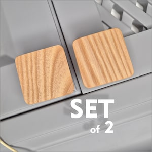 May include: Two square wooden coasters with rounded corners, featuring a natural wood grain pattern. The coasters are light brown and are set against a gray background. The text "SET of 2" is displayed in white.