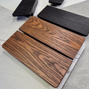 Op de afbeelding: Houten lade- en kastgrepen in verschillende afwerkingen. De afbeelding toont rechthoekige grepen in donkerbruine, zwarte en natuurlijke houttinten. De houtnerf is zichtbaar op de natuurlijke houten stukken.