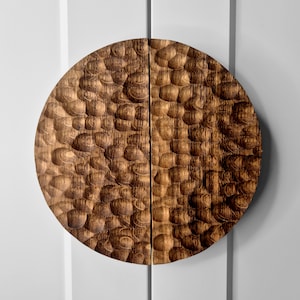 May include: A circular wooden wall art piece composed of two semi-circular halves. The surface features a deeply carved, textured pattern of overlapping, rounded indentations that create a rhythmic, scalloped appearance across the brown wood grain.
